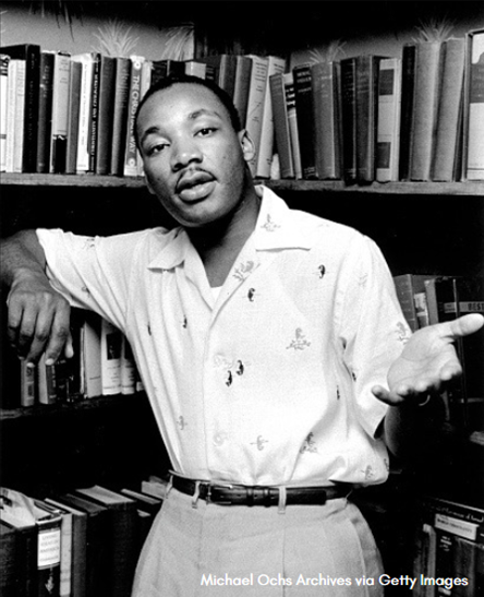 Black and white photo of Martin Luther King with his hand outstretched standing in front of books on shelves